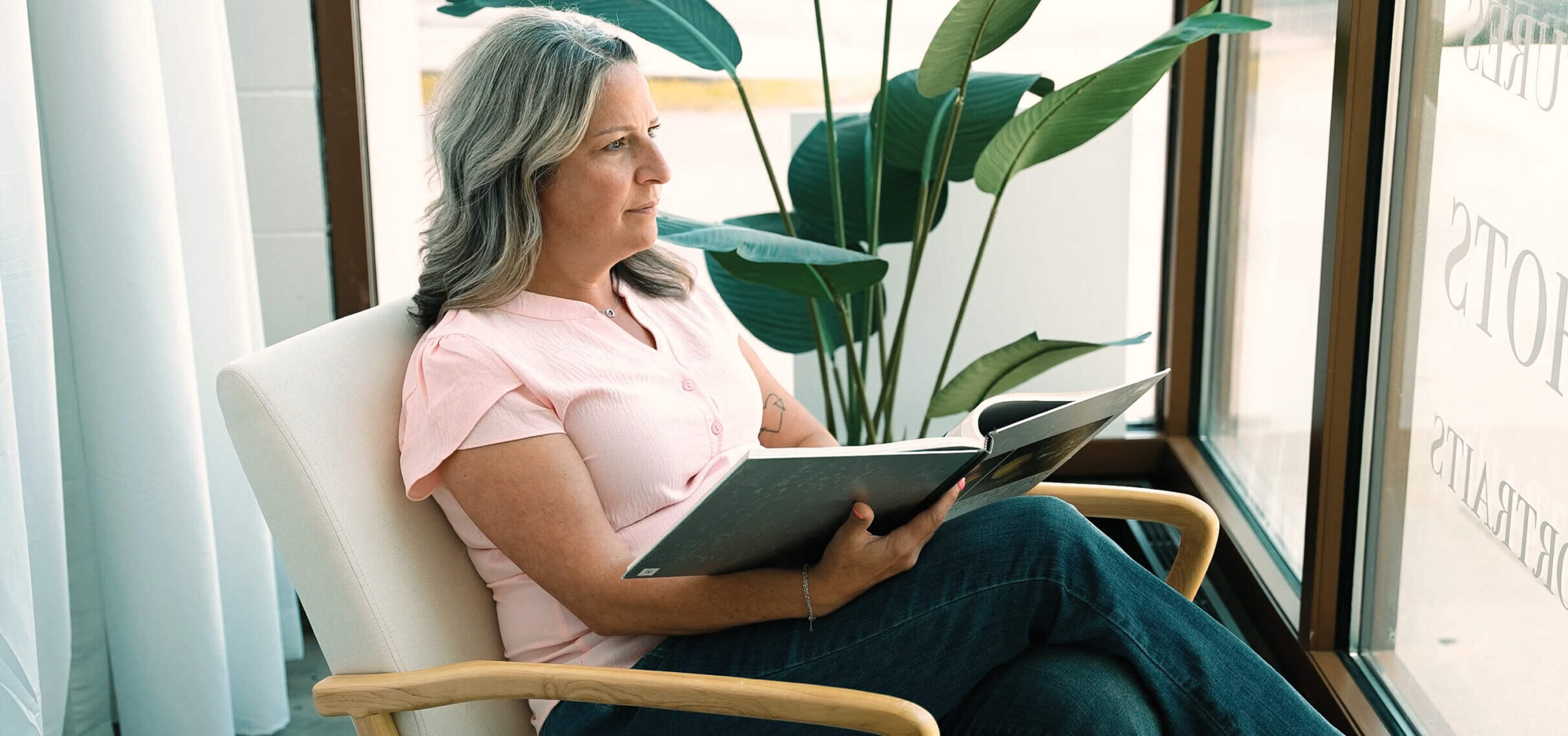 Kristin Miller, a breast cancer survivor and mammography tech at Envision Radiology sits by a window with a book in her lap.